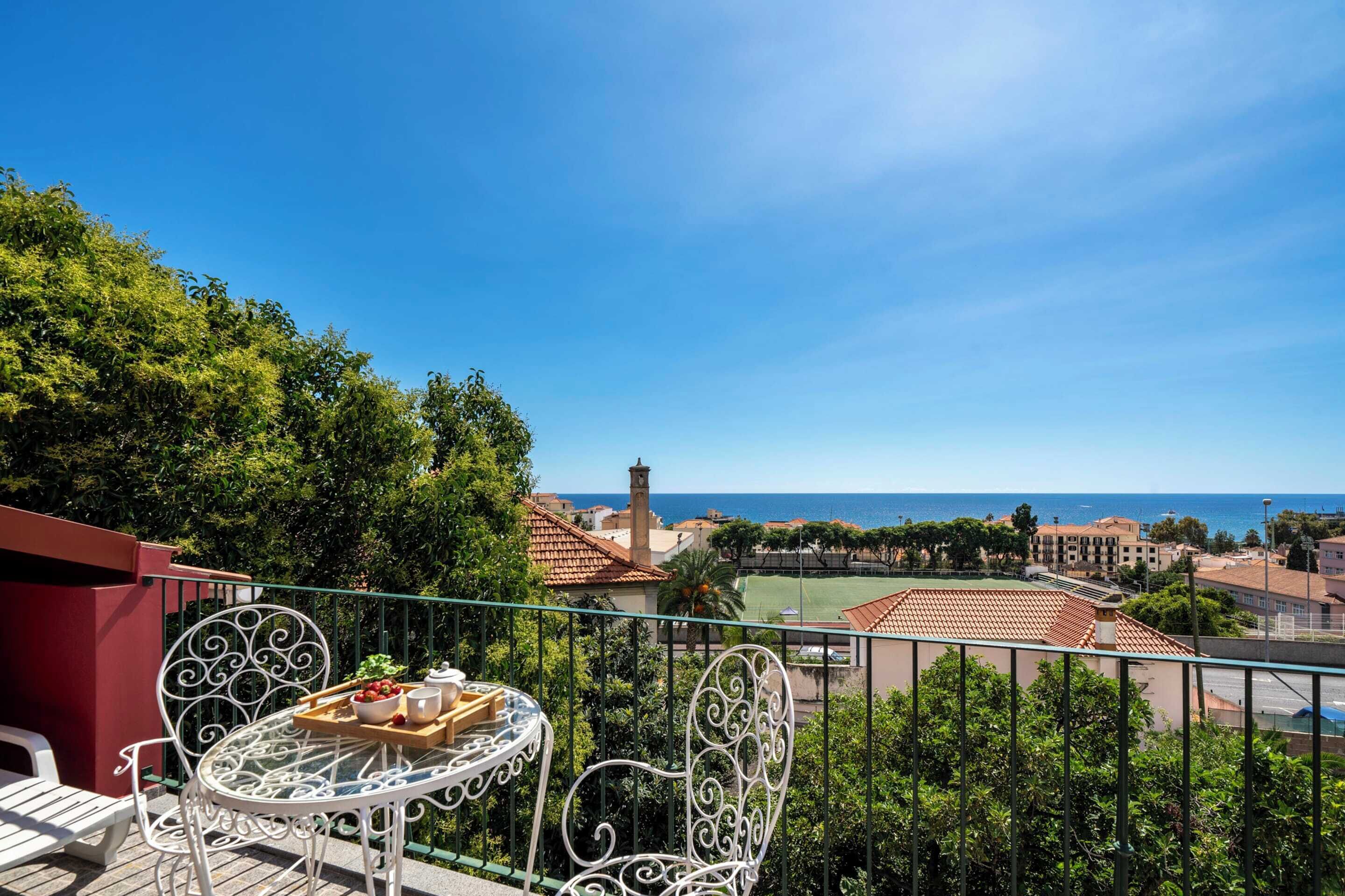 Un balcón con mesa y sillas, con vista a un pueblo costero con edificios y al mar.