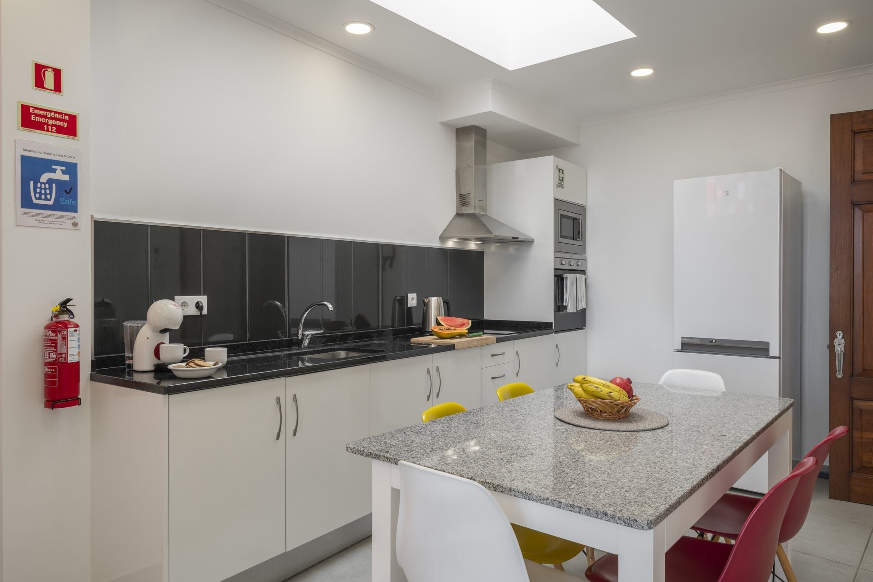A modern kitchen with a granite countertop, white cabinets, and a stainless steel range hood. There is a fire extinguisher mounted on the wall, and a small dining area with a table and chairs.