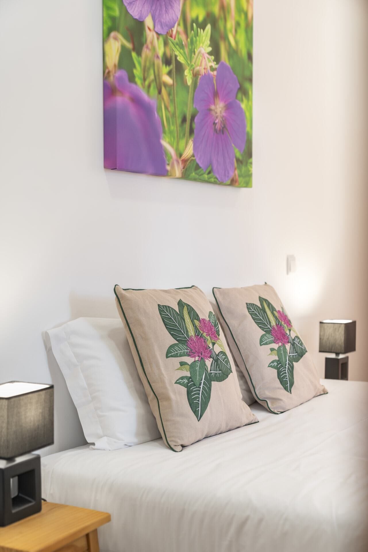 Un dormitorio con cama, dos almohadas con bordado floral, una lámpara de noche y un cuadro de flores colgado en la pared.