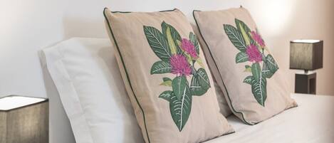 Un dormitorio con cama, dos almohadas con bordado floral, una lámpara de noche y un cuadro de flores colgado en la pared.