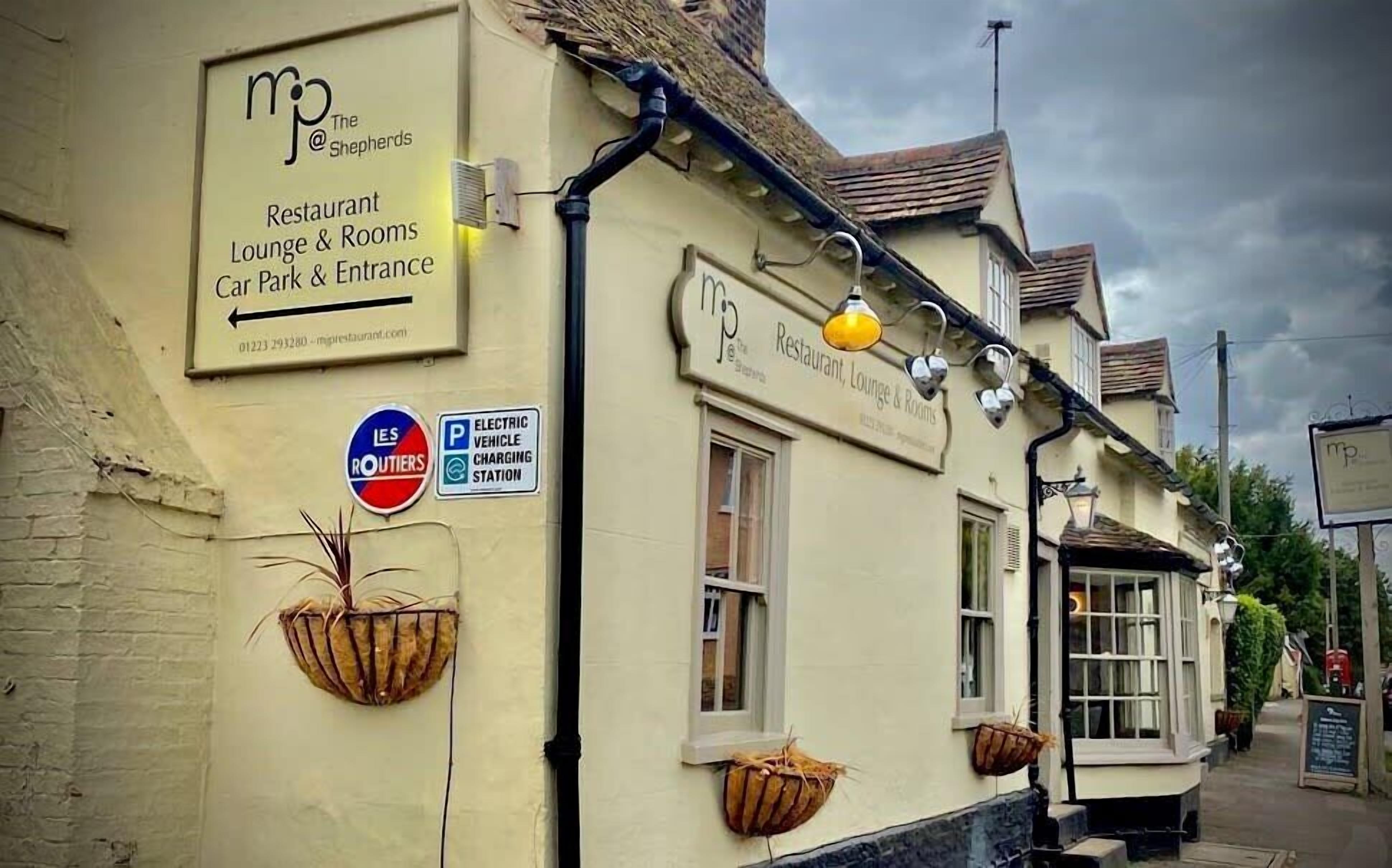 Un edificio con un cartello che indica il ristorante, il lounge bar e le camere 'The Shepherds'. Segnale che indica la presenza di una stazione di ricarica per veicoli elettrici nelle vicinanze.