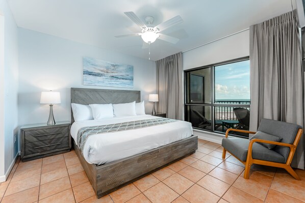 A hotel room with a large bed, two bedside tables, a ceiling fan, a view of the outdoors, and a chair.