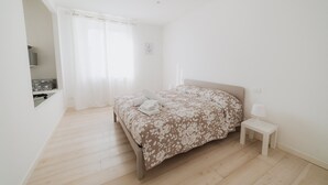 Un dormitori amb un llit, una taula de nit, una petita prestatgeria i una finestra amb cortines.