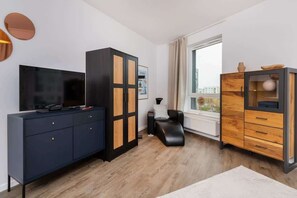 A modern living room with a flat-screen TV, dark blue and wooden cabinets, a black leather chair, and a large window with a view of buildings.