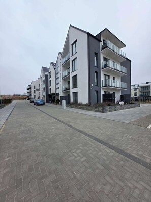 A modern apartment building with a brick-paved walkway, multiple balconies, and a clear sky.