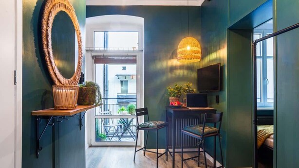 A modern interior with a large mirror, a hanging light fixture, a small table with a chair, and a bed in the background. - Cozy Studio in the Center | Balcony | Close to Train Station (Katowice)