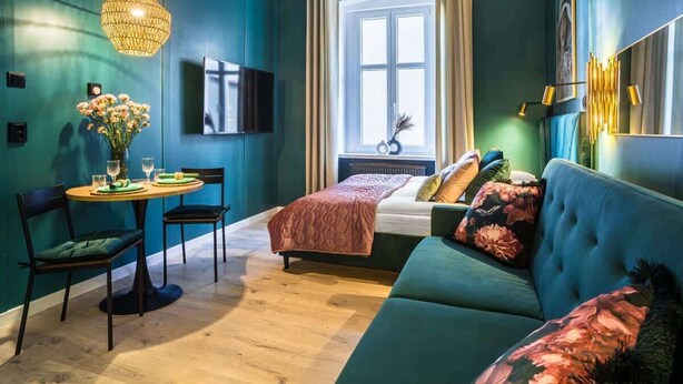 A modern room with teal walls, a wooden dining table with chairs, a sofa, a bed with a red blanket, and a large window with curtains.