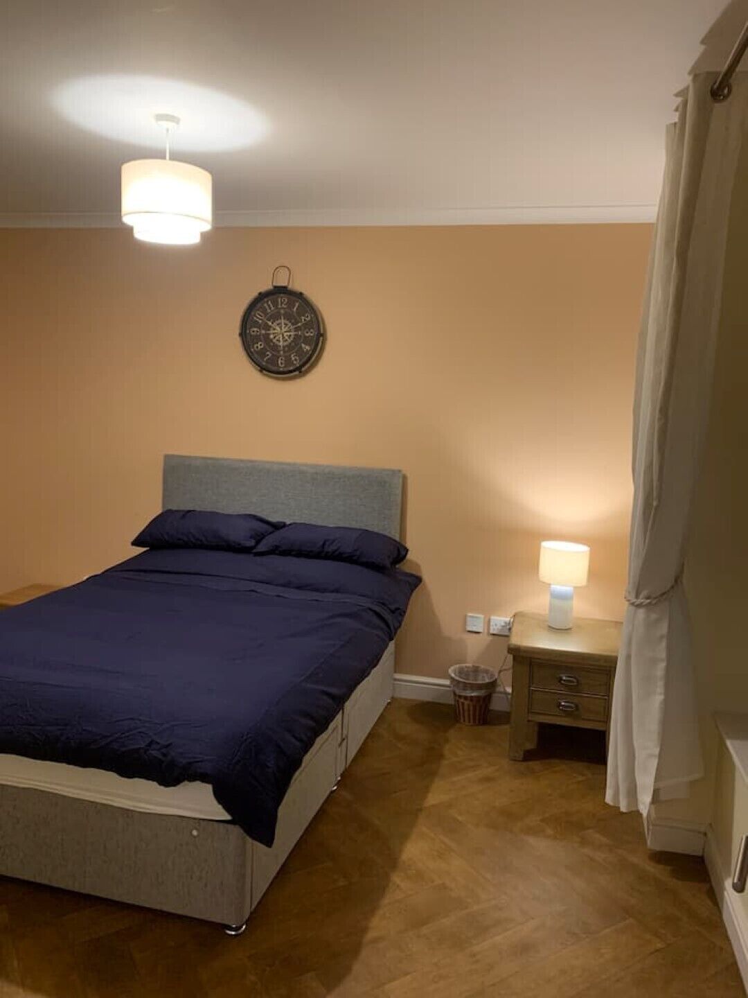 Et soveværelse med seng, natbord, lampe og et ur på væggen.