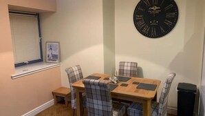 Un área de comedor con una mesa de madera, sillas a cuadros, un reloj de pared grande, una ventana con persianas y una mesita auxiliar.