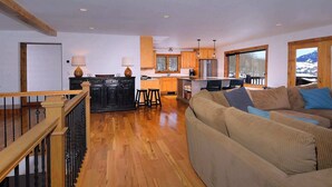 Amplia sala de estar con piso de madera, un sofá, una cocina con alacenas de madera y vistas al exterior.
