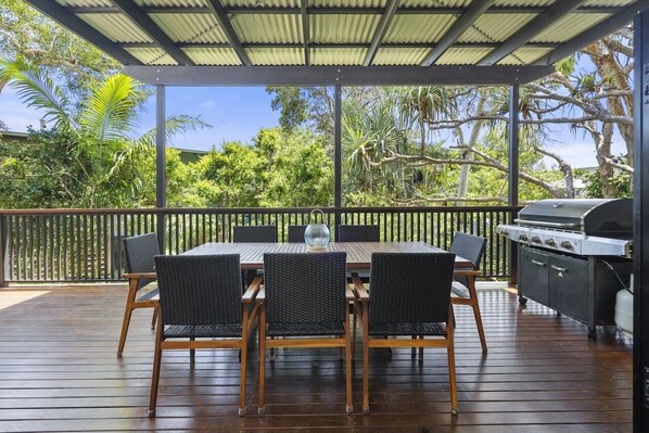An outdoor patio with a wooden table and chairs, a grill, and a view of tropical trees.