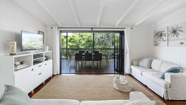 Un salón amplio con un sofá blanco, un televisor sobre un mueble blanco, una zona de comedor con mesa y sillas, y un balcón con vistas a la vegetación.