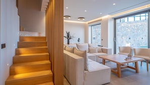 Een modern interieur met een houten trap, een zithoek met banken en een tafel, en grote ramen met uitzicht op een stenen gebouw.