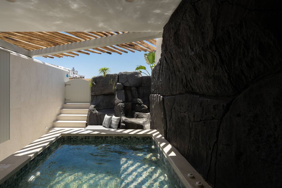 Piscina com borda revestida em azulejo, pergolado de madeira acima e parede de pedra.