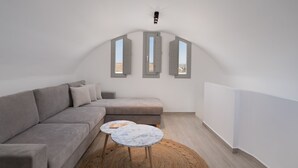 En moderne stue med en grå sofa, et rundt bord og tre vinduer med skodder.