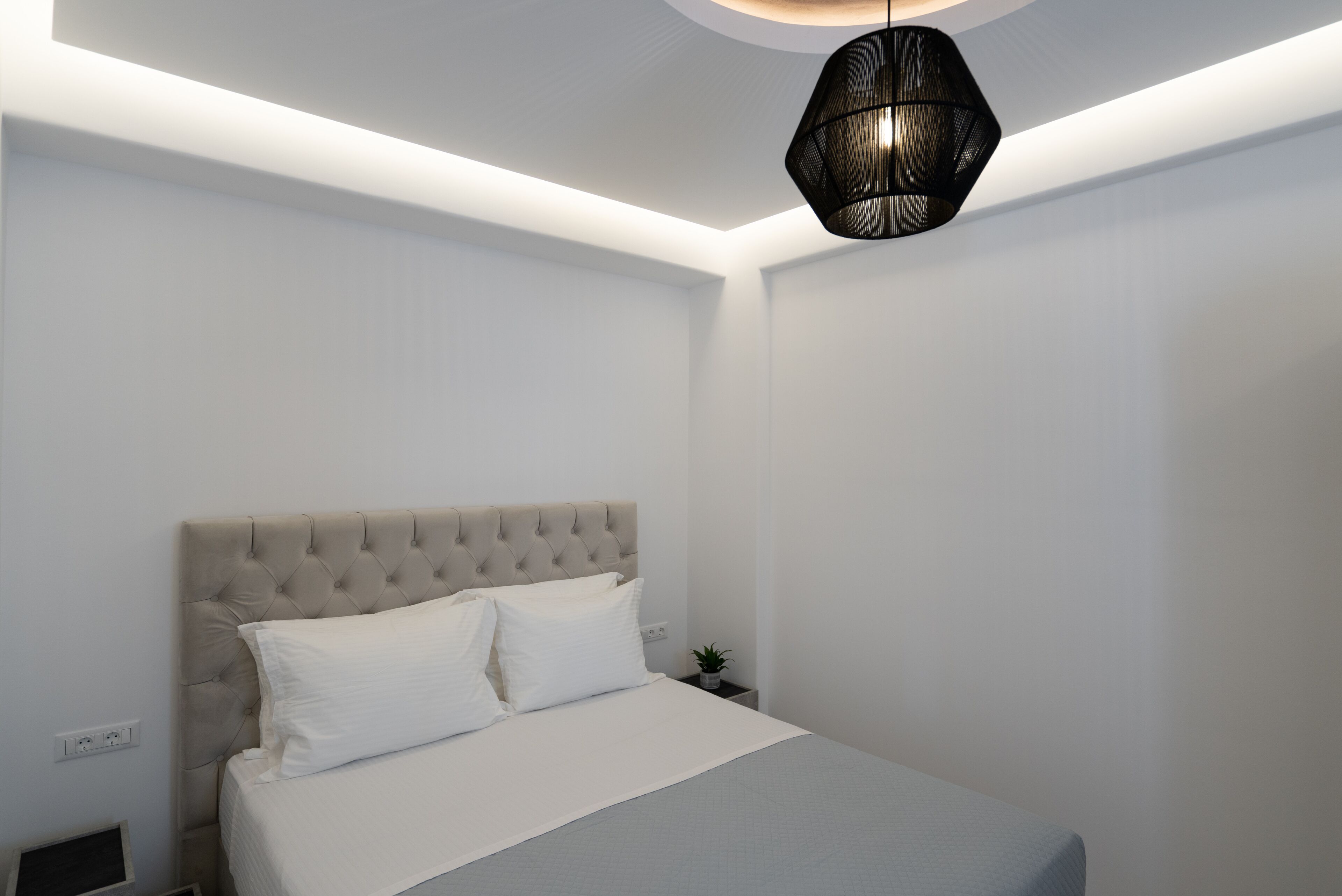 A bedroom with a tufted headboard, a bed with white linens, a small bedside table with a plant, and a modern pendant light.