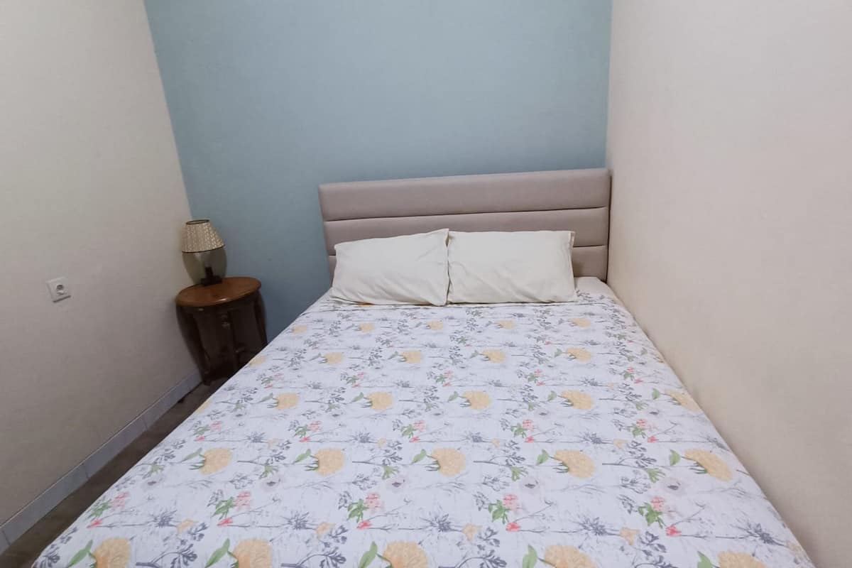 Ein kleines, ordentlich bezogenes Bett mit floraler Tagesdecke, ein hölzerner Nachttisch mit Lampe und eine hellblaue Wand.