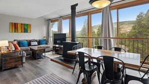A modern living room with a sofa, coffee table, TV, and fireplace. Large windows offer a view of the outdoors.