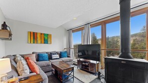 A living room with a sofa, coffee table, TV, fireplace, and large windows offering a view of the outdoors.