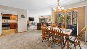 A spacious living area with a dining table, chairs, and a chandelier. There is a kitchen area with wooden cabinets and a large window overlooking a snowy landscape. - Nostalgic Mountain Getaway, Only a Short Walk to Lifts or River Run! (Keystone)