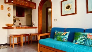 A cozy living area with a wooden bench, a blue sofa with tropical cushions, a kitchen area with a wooden counter and stools, and a framed picture of a sunflower on the wall.