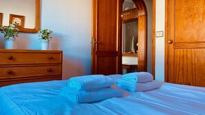A neatly made bed with blue linens, a wooden dresser with drawers, a potted plant, and a wooden door with a mirror.