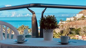 A table with two cups and saucers, a potted plant, and a view of the sea and buildings.