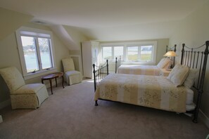 A bedroom with two beds, a chair, a table, and large windows offering a view of the outdoors.