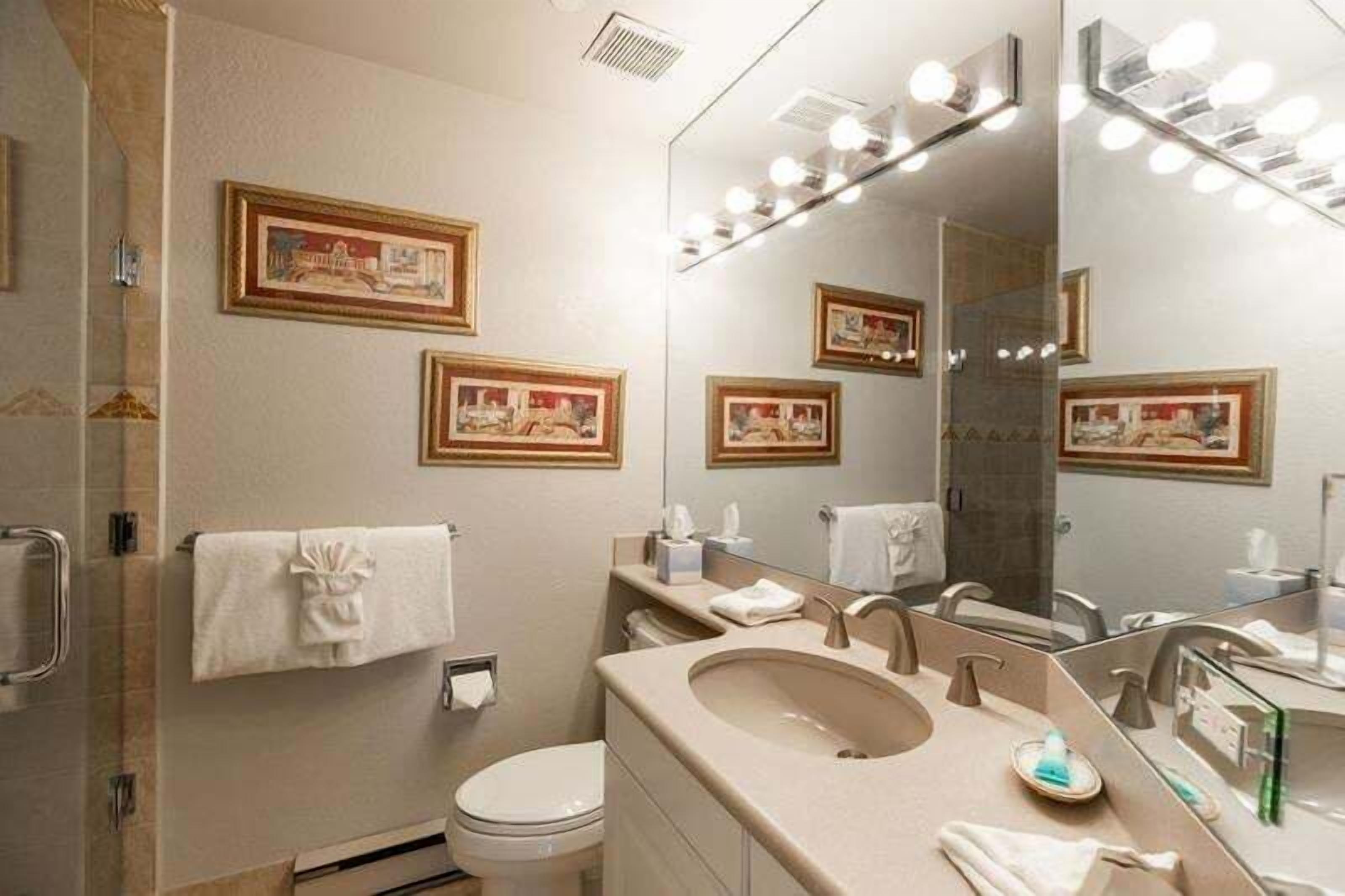 A bathroom with a large mirror, framed pictures on the wall, a sink, and a toilet.