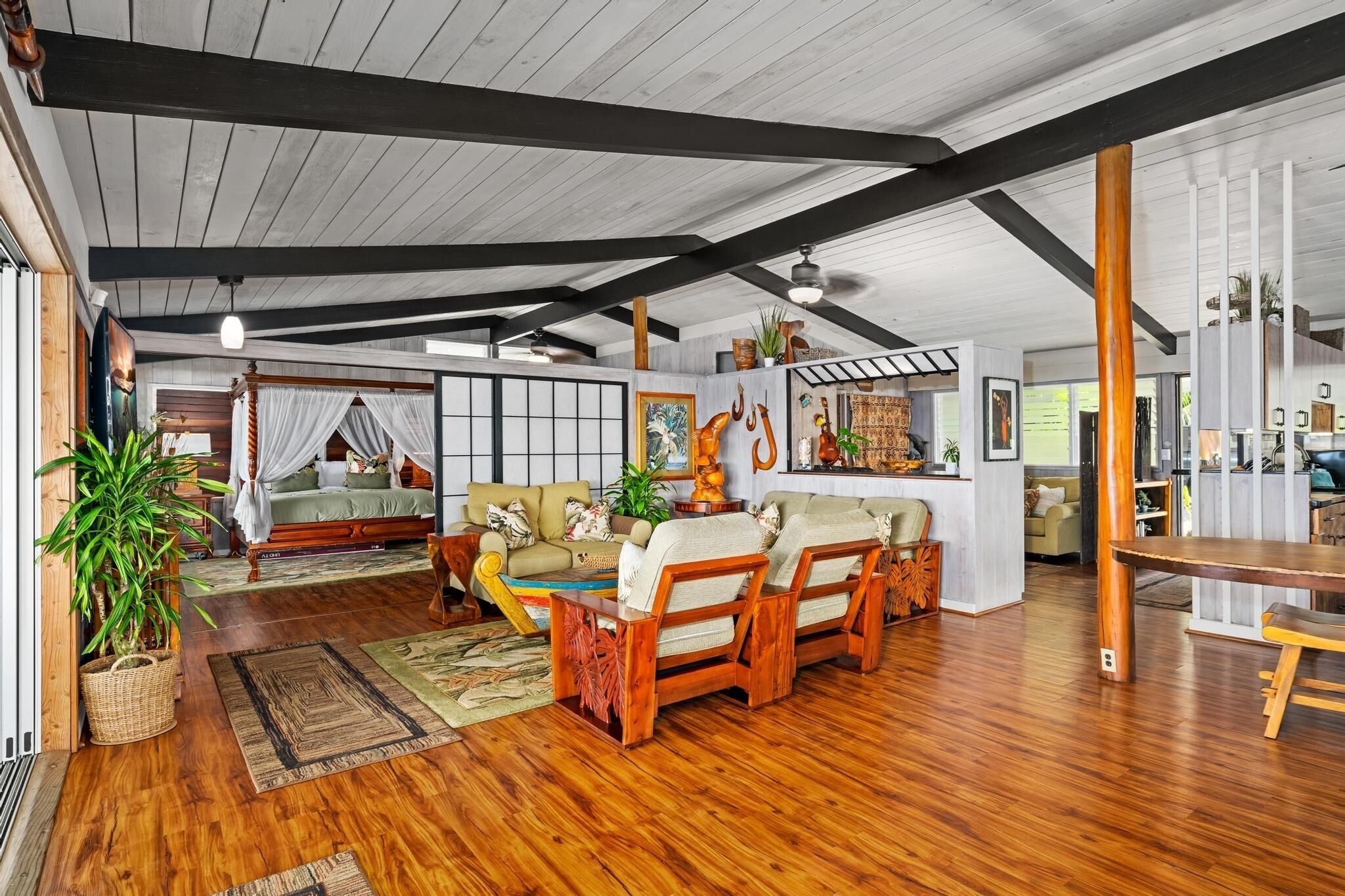 A spacious living room with a large sofa, a wooden coffee table, and a dining table. There is a bed with a canopy in the background, and a kitchen area is visible to the right.