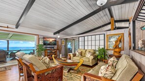 A spacious living room with wooden furniture, a large window overlooking the ocean, and a view of the sea.