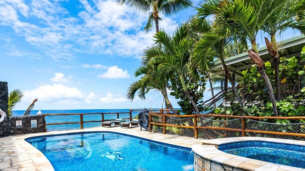A swimming pool with clear blue water, surrounded by stone walls and palm trees, with a view of the ocean in the background.