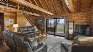 A log cabin interior with a wooden ceiling, leather sofas, a TV, and a large window.
