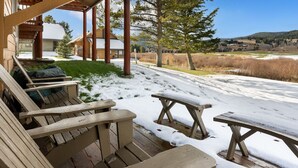 Une terrasse en bois, dotée de bancs et d’une table de pique-nique recouverts de neige, offre une vue sur un paysage rural avec des arbres et, au loin, sur des maisons.