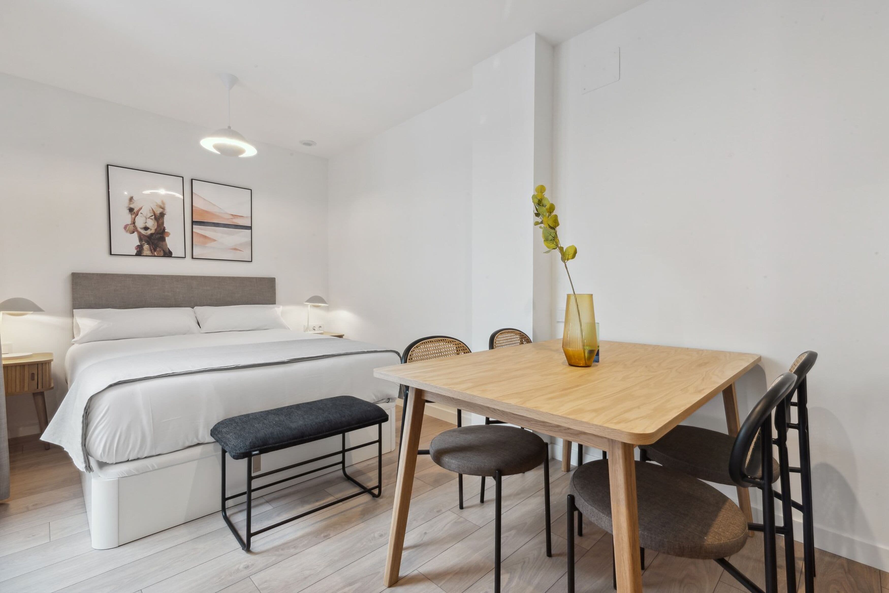 Un dormitorio moderno con una cama grande, mesitas de noche, una lámpara de noche, una mesa de comedor de madera con sillas y un florero con flores.
