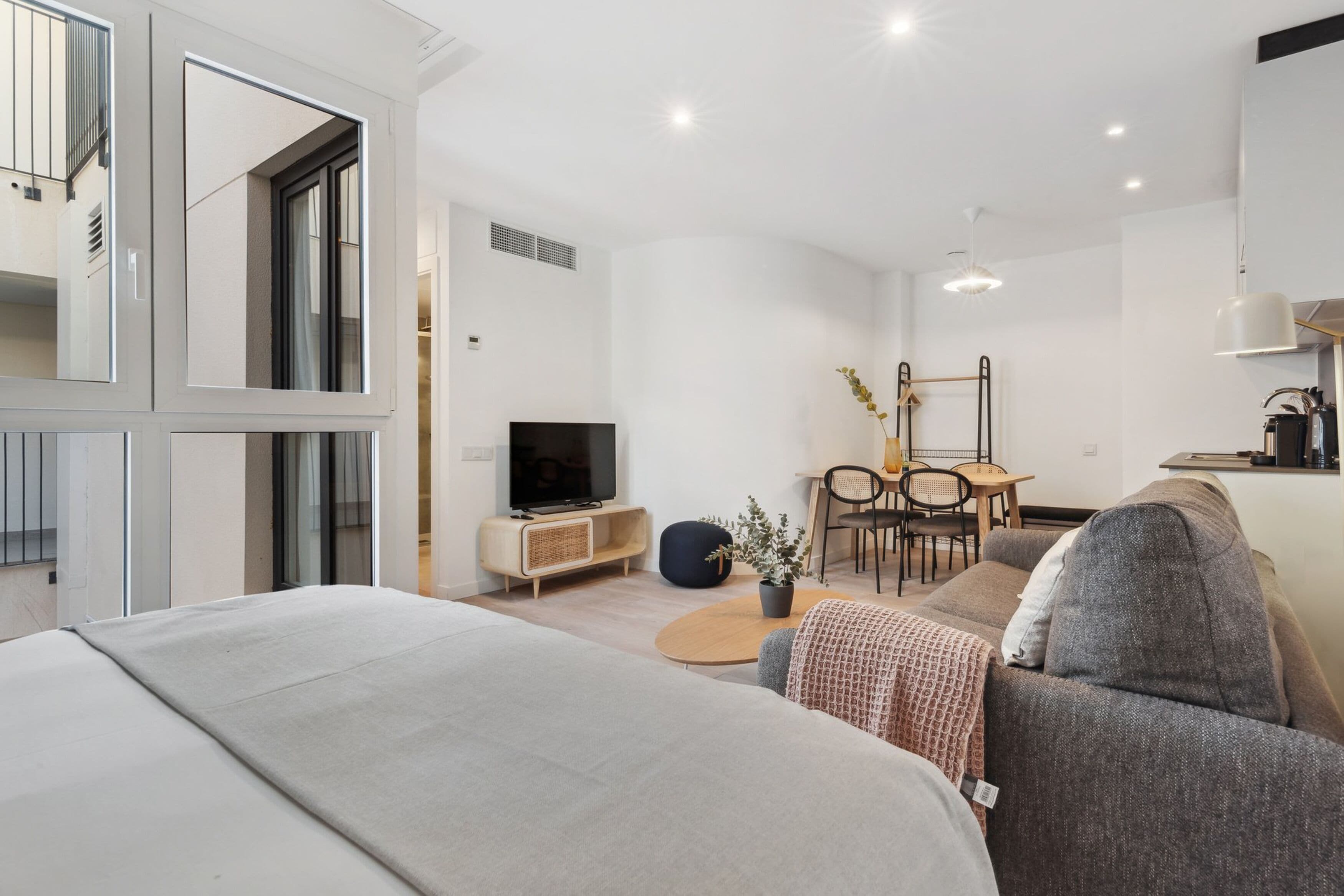 A modern, well-lit living room with a large bed, a TV, a dining area, and a kitchen.