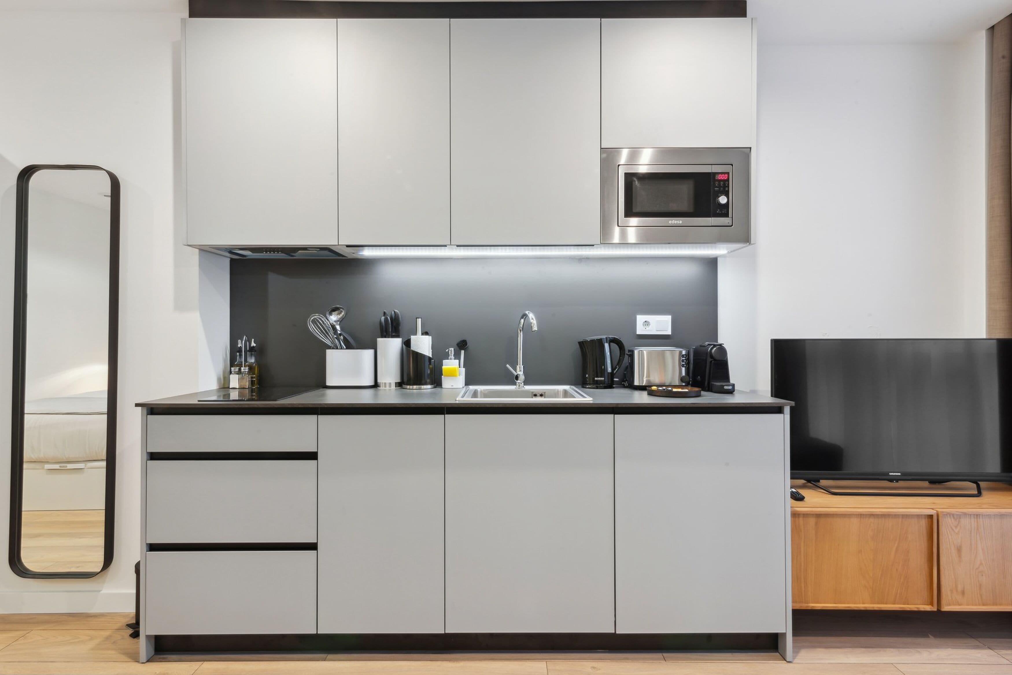Une cuisine moderne avec des meubles gris, un four à micro-ondes en acier inoxydable, un évier et un téléviseur à écran plat.