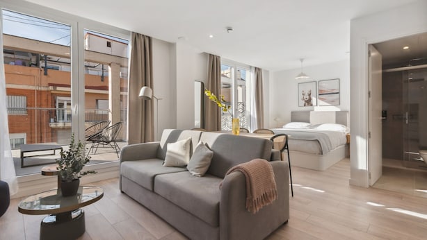 Una sala de estar moderna con un sofá gris, una mesa de centro redonda de vidrio y una cama al fondo.