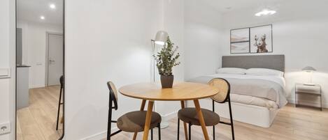 A modern, well-lit room with a bed, bedside table, lamp, and a dining area with chairs and a table.