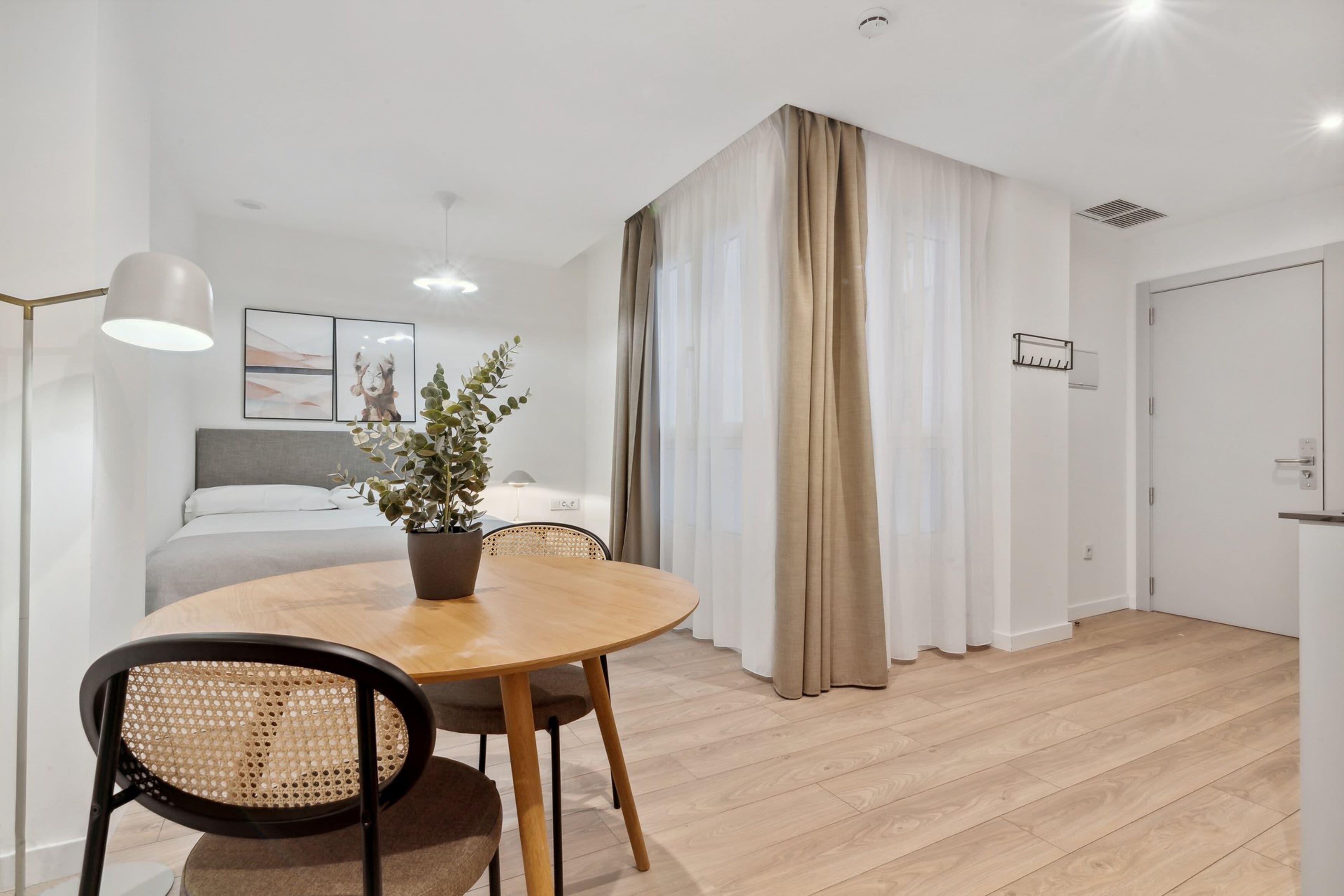 Una habitación moderna y bien iluminada con una mesa de comedor redonda de madera, dos sillas, una planta en maceta y una cama con cabecero gris.