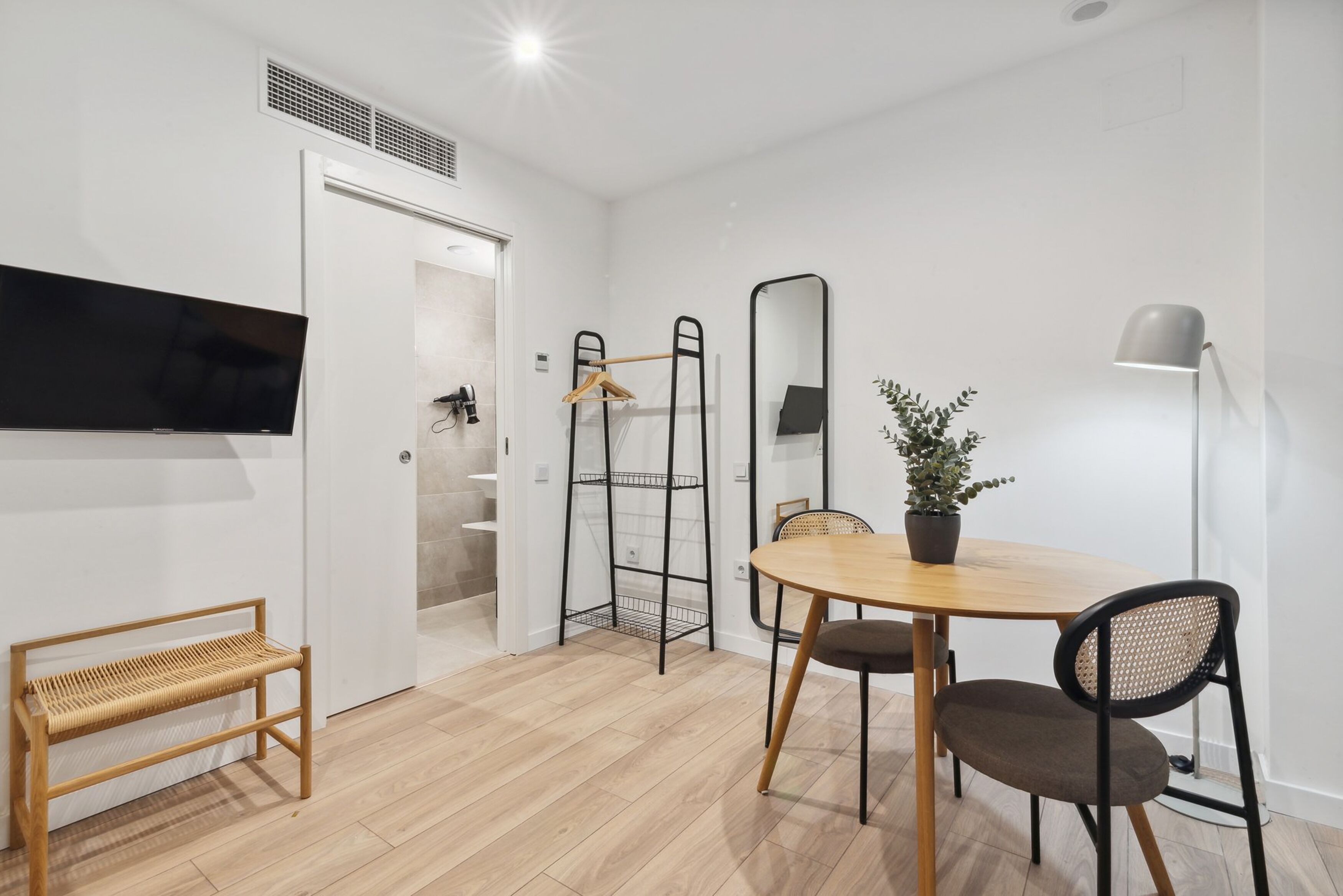 Una habitación moderna con suelo de madera, una mesa redonda con sillas, un televisor montado en la pared, un perchero, un espejo y una planta en maceta.