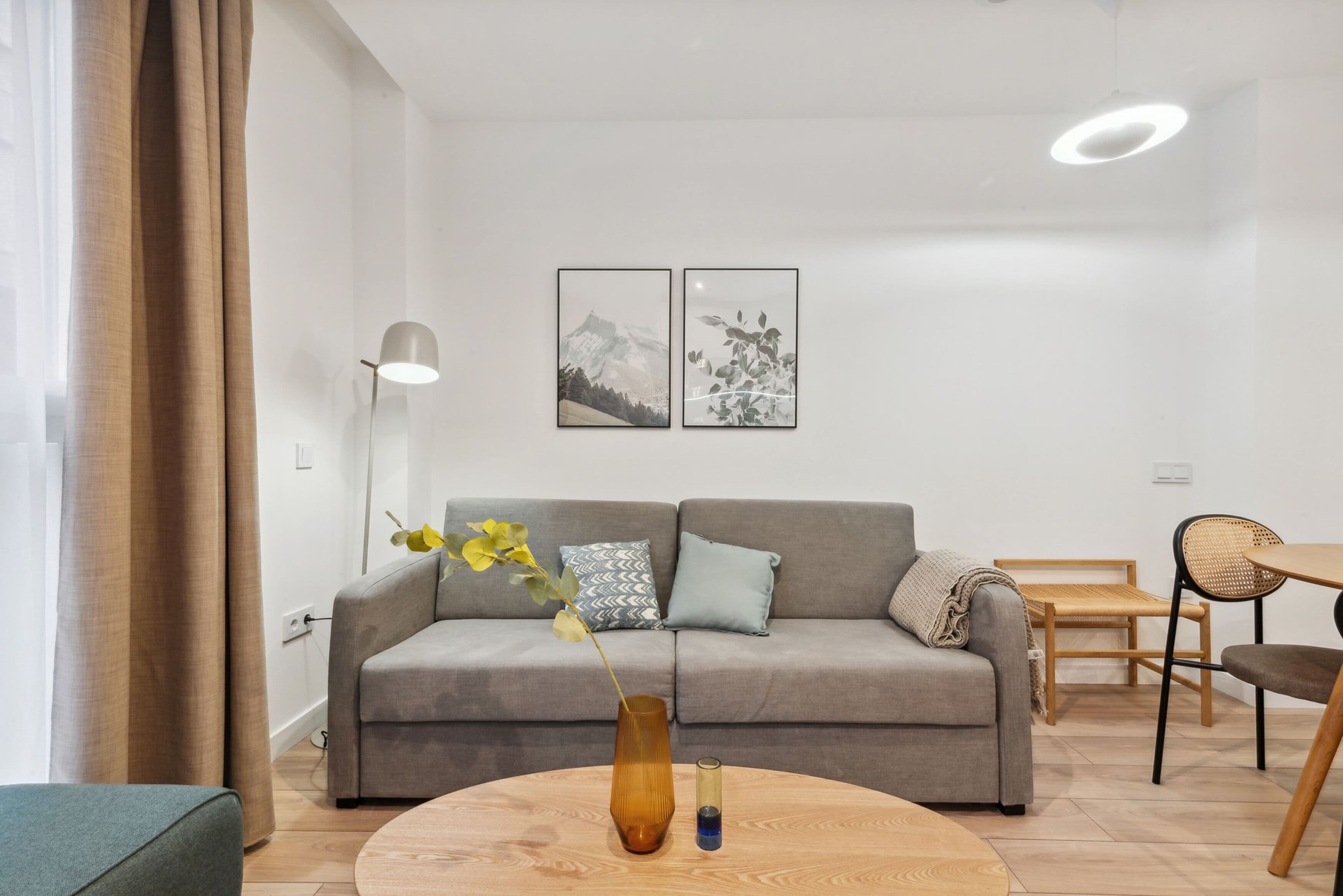 Un salon moderne avec un canapé gris, une table basse ronde en bois et un vase rempli de fleurs. Deux tableaux encadrés ornent le mur, ainsi qu’une lampe et un coin repas avec une table et des chaises.