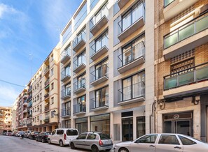 Un edificio residencial de varios pisos con balcones, una tienda en la planta baja y coches aparcados a lo largo de la calle.