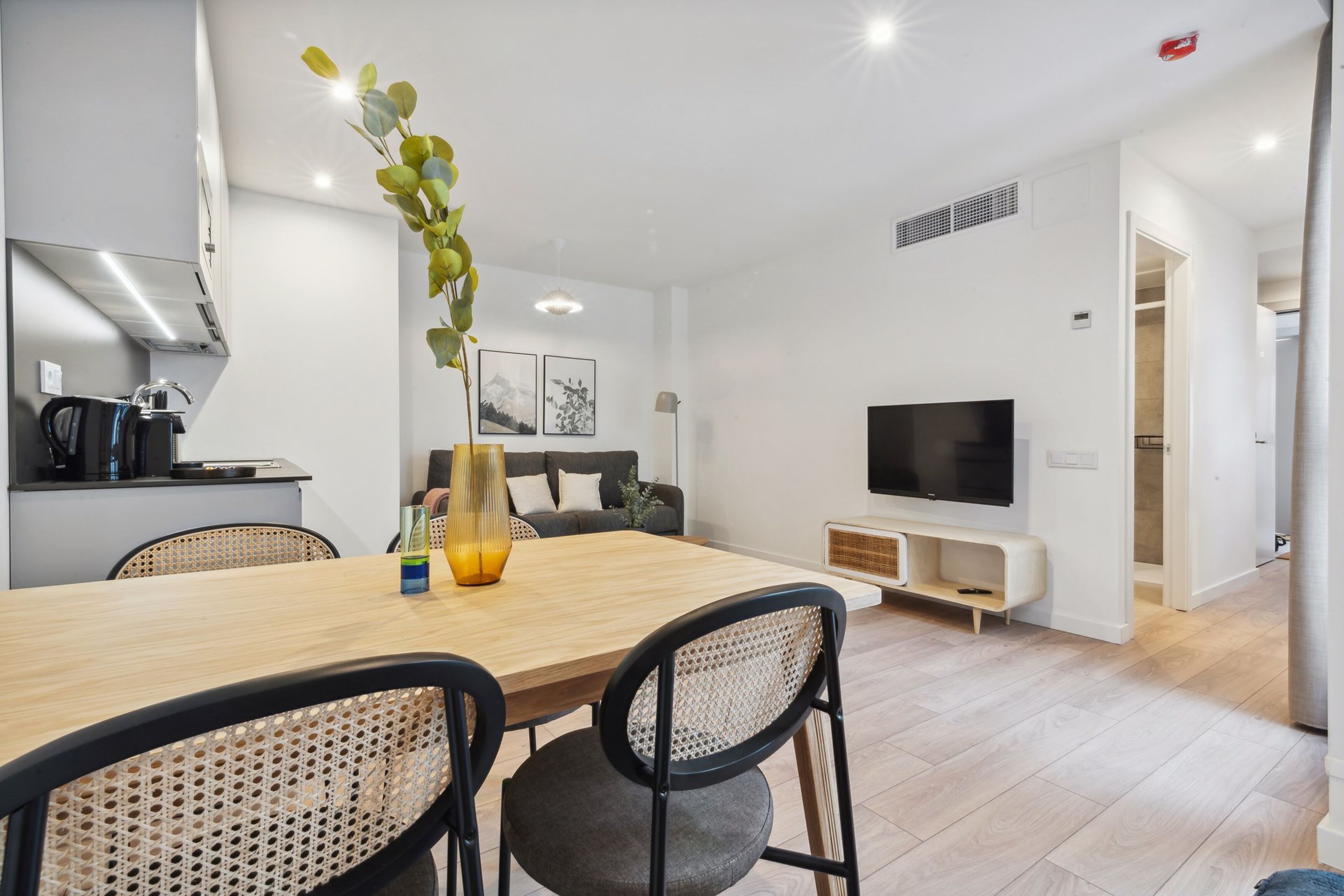 A modern living room with a wooden dining table, black and white chairs, a sofa, a TV, and a kitchen area with a kettle.