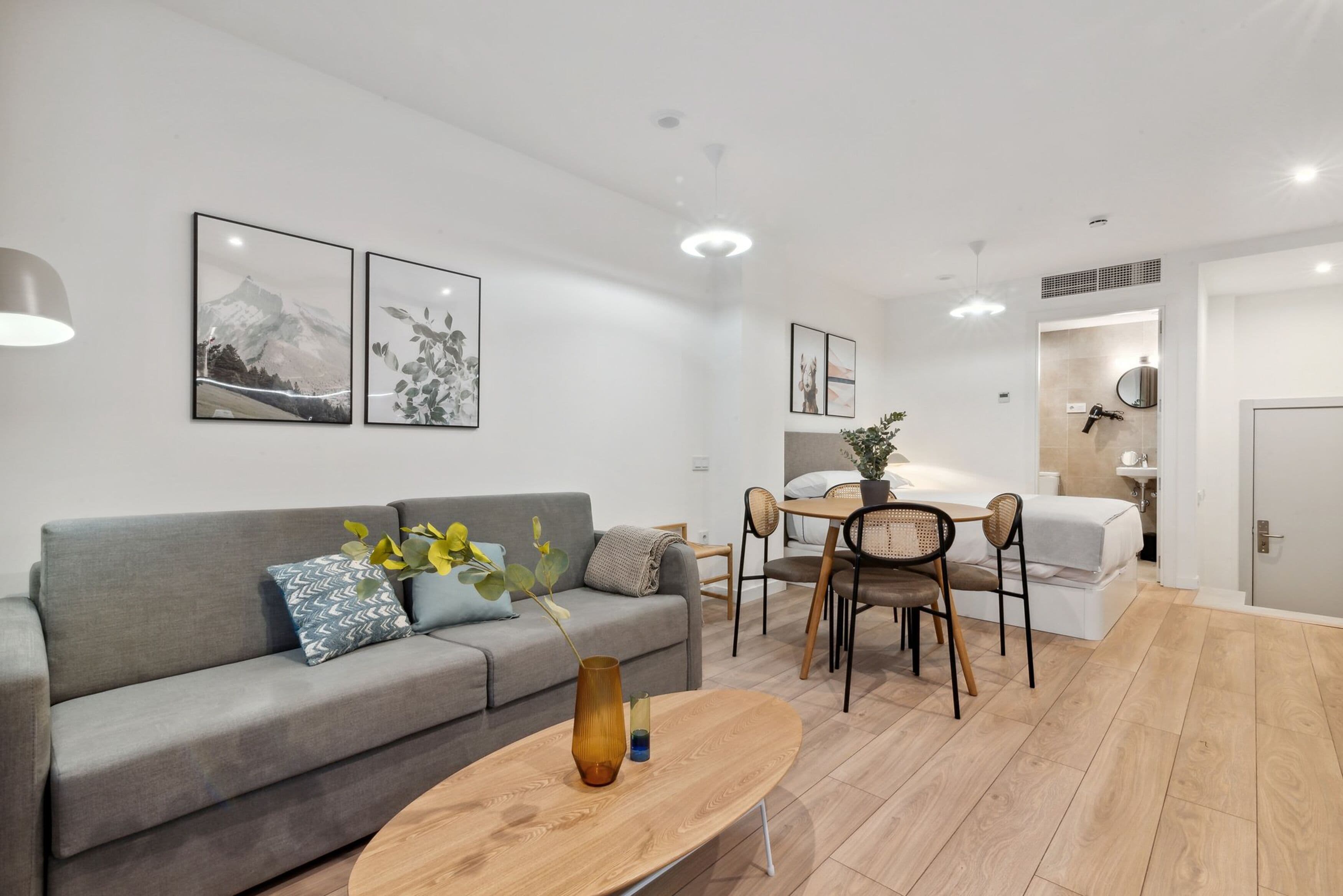 A modern living room with a grey sofa, a wooden coffee table, and a dining area with a table and chairs. There are framed pictures on the wall and a ceiling light fixture.