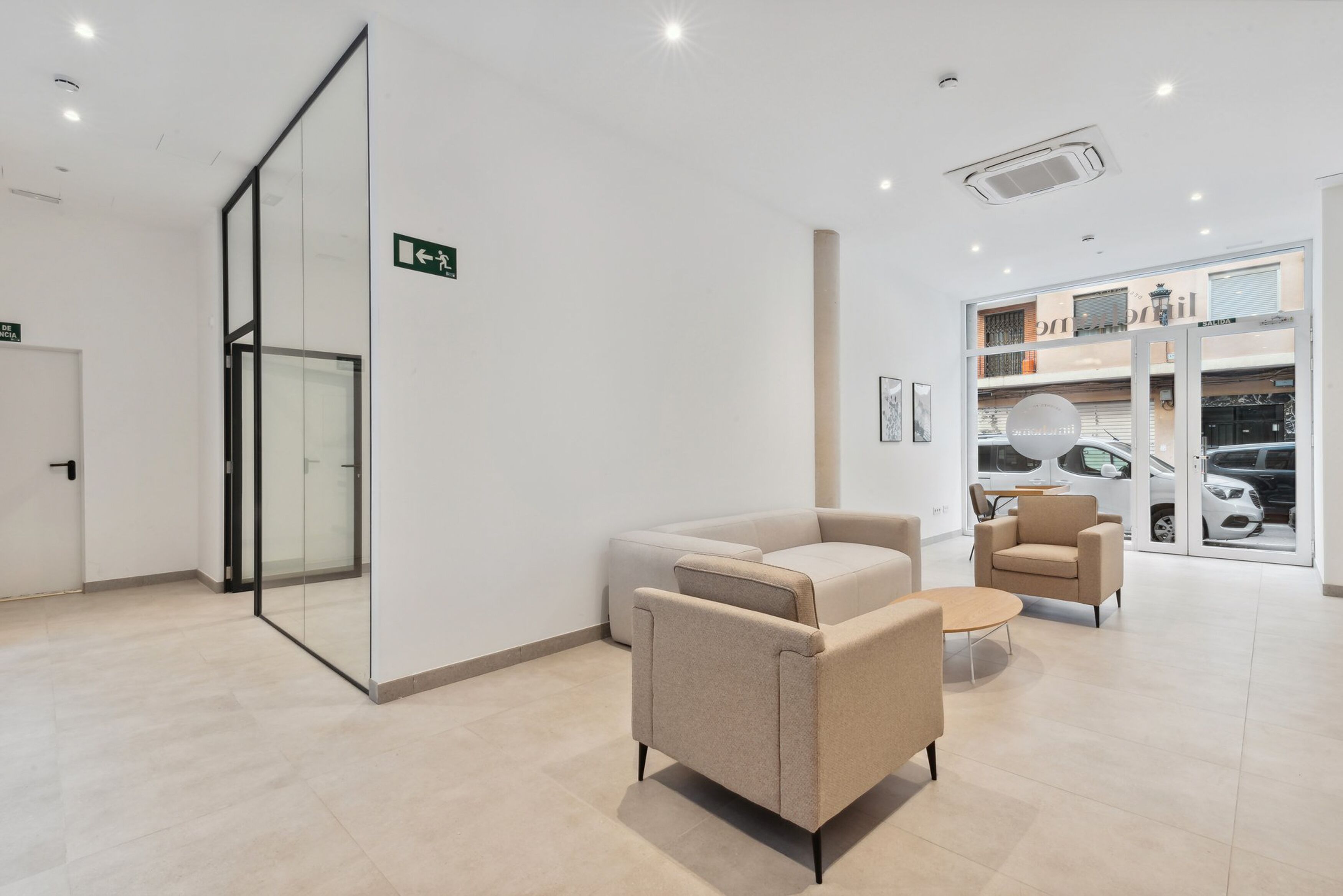 A modern waiting area with a glass partition, beige sofas, and a view of parked cars through the glass doors.