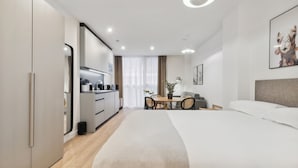 Una habitación de hotel moderna con una cama grande, un área de comedor con mesa y sillas, una kitchenette y un ventanal con cortinas.