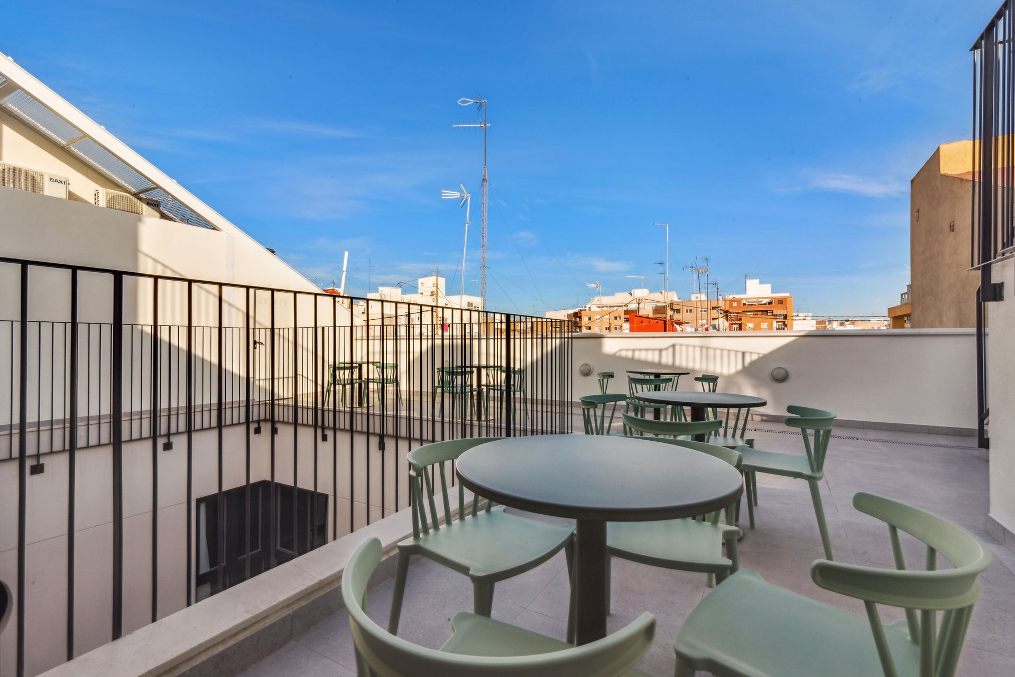 A rooftop terrace with round tables and green chairs, surrounded by a black metal fence.
