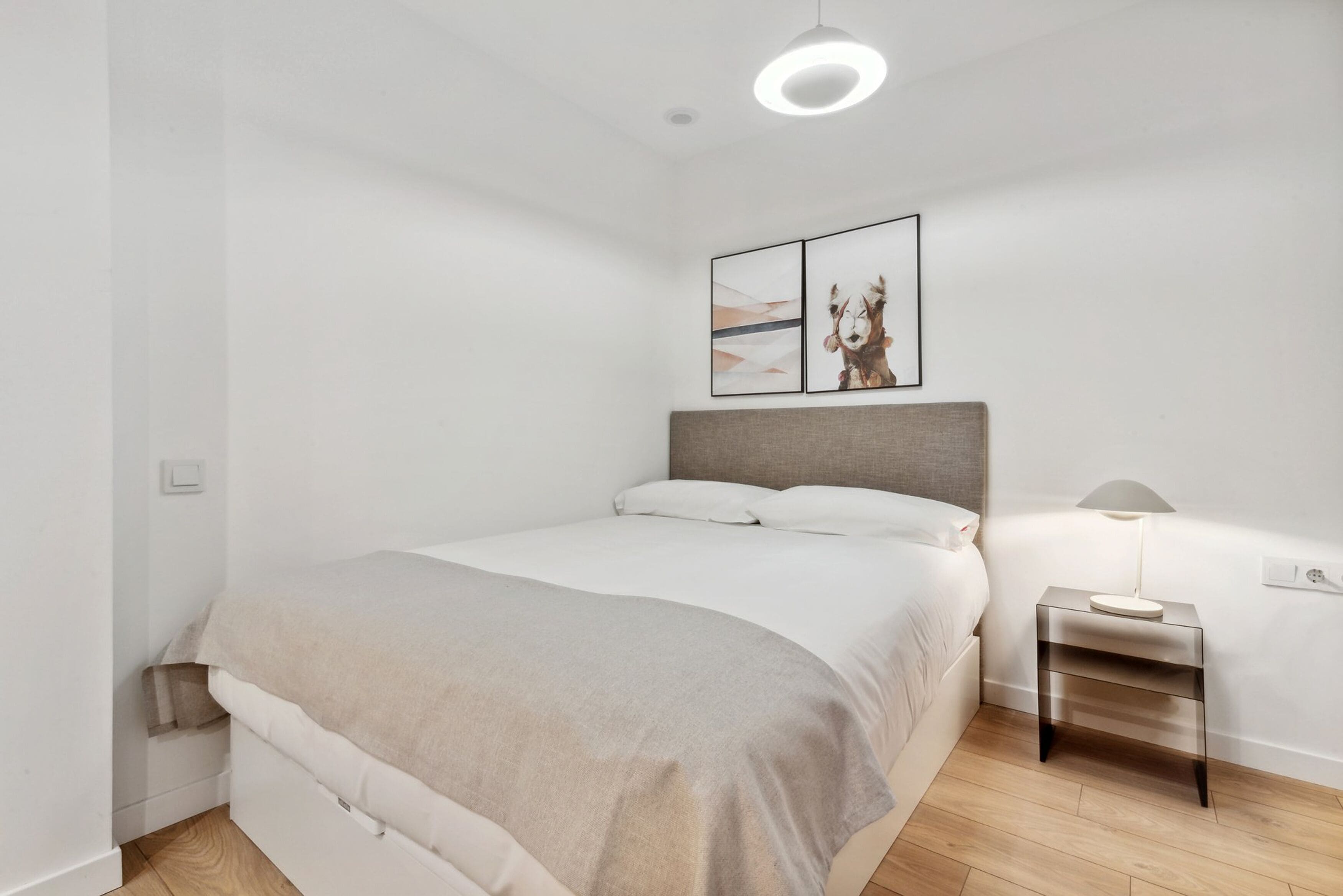 Un dormitorio minimalista con una cama grande, una mesita de noche con lámpara y un cuadro en la pared.
