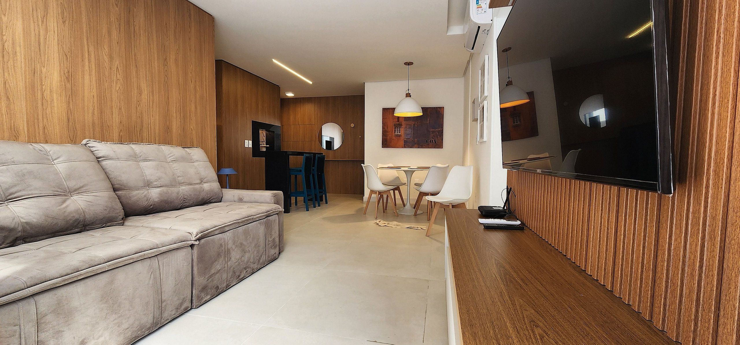 Uma sala de estar moderna com um sofá, um espaço de refeições com cadeiras e uma TV fixada na parede.